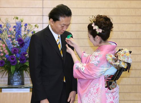 Photograph of the Prime Minister having a green feather pin attached by the Japan Cherry Blossom Queen